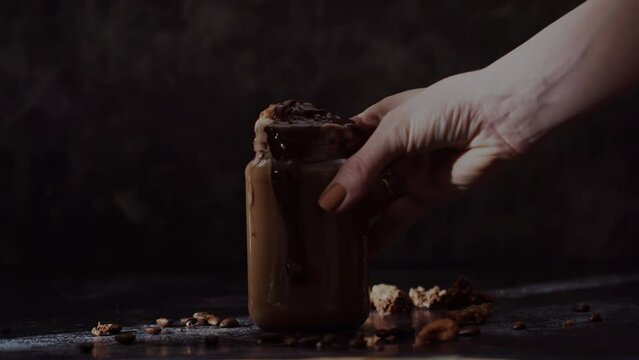 Female Hand put Ice Coffee on moody background in slow motion. Delicious decorated glas of iced coffee topped with cream, ice cream and chocolate pieces, cookies and chocolate sauce.