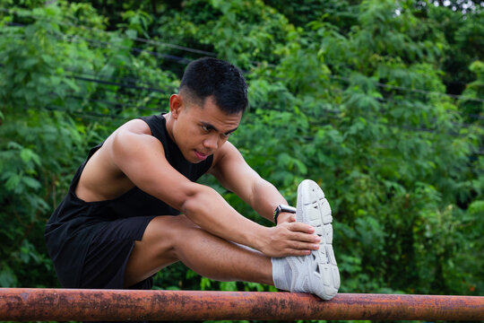 Athletic Filipino man on sports clothes stretching leg on railing before workout at park. Outdoors activity, exercise warm up, healthy lifestyle concepts