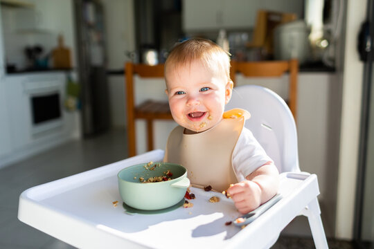 Happy seven month old baby eating breakfast in highchair in morning sunlight