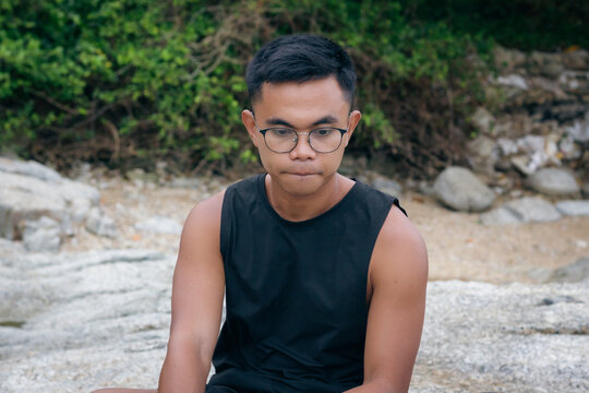 Pensive Filipino man with eyeglasses looking nervous sitting alone on rocks. Worried male adult with thoughtful look, solitude concepts