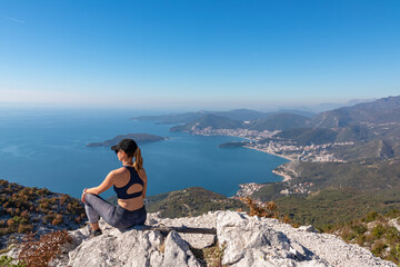 Hiking woman sitting on rock enjoying panoramic aerial view on coastline of Budva and Sveti Nikola Island seen from Goli Vrh, Adriatic Mediterranean Sea, Montenegro, Balkan, Europe. Budvanian Riviera