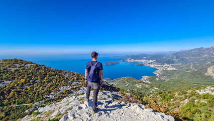 Naklejka premium Hiking man with backpack enjoying panoramic aerial view on coastline of Budva and Sveti Nikola Island seen from Goli Vrh, Adriatic Mediterranean Sea, Montenegro, Balkan, Europe. Budvanian Riviera