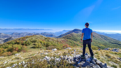 Naklejka premium Man with backpack and hat on top of Goli Vrh with scenic view on mountain chains of Dinaric Alps and Lake Skadar National Park, Montenegro, Balkan, Europe. Valley covered by mystical fog. Freedom