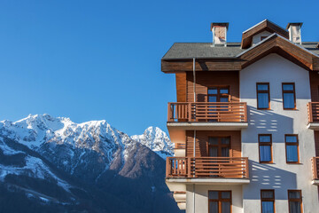 Multi-storey wooden residential building with sloping roofs in the European style against the backdrop of a mountain snow-capped peak with forests and blue sky. - obrazy, fototapety, plakaty