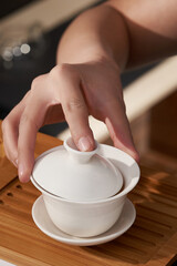 Shot of female hands holding a white original porcelain color printed gaiwan in chinese style. The designer bowl with a lid and a saucer is located on a wooden tray on a dark background.