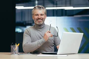 Portrait of senior man inside office, mature businessman with beard smiling and looking at camera, boss working at desk using laptop at work.