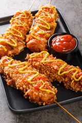 Korean corn dogs are made from either sausage, mozzarella cheese on skewer and coat this in a sweet flour batter before rolling in a layer of toppings closeup on the plate on the table. Vertical
