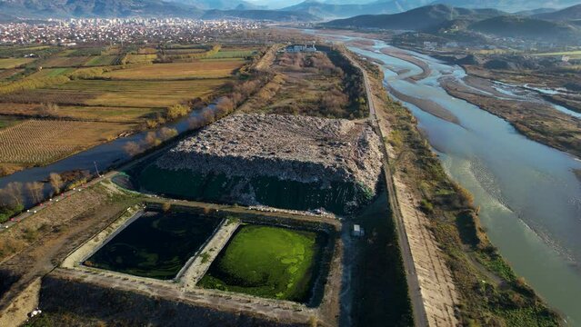 Landfill with huge waste mountain near the river and agricultural land with Elbasan cityscape background in Albania