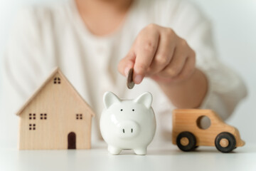 Close up of woman hand putting coin into White piggy bank for saving money, toy house and car on...