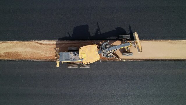 Road construction grader aerial view. Construction grader levels the ground between an asphalt road for further installation of road fenders. Road construction works. Heavy machinery. Aerial footage