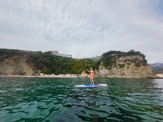 Fit woman in bikini relaxing on a sup surfboard, floating on the clear sea on cliff background