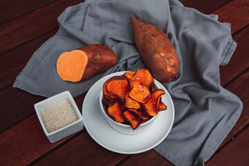 Sweet potato chips with sesame seeds in a white ceramic bowl. With a grey kitchen cloth in the background.