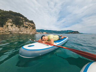 Fit woman in bikini relaxing on a sup surfboard, floating on the clear sea on cliff background