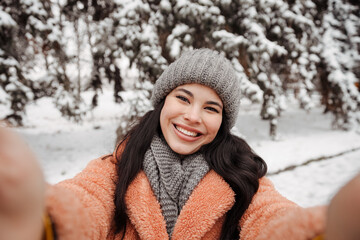 Pretty happy woman in gray hat and pink coat takes selfie and play the ape in winter cold day. Girl takes winter selfie