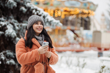 Pretty long-haired girl with smartphone at winter snow park in cold weather scrolling texting chatting using mobile phone