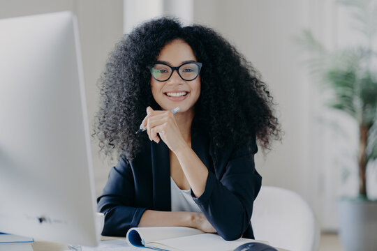 Positive International Female Student With Crisp Hair, Wears Transparent Glasses, Holds Pen In Hand, Makes Accountings, Sits In Front Of Big Computer Screen, Dressed In Black Elegant Outfit.