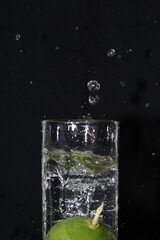 Lima verde cayendo en el vaso de agua de vidrio transparente produce  burbujas en el aire, formando un original y atractivo diseño con fondo negro