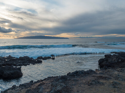 View of the natural sea rock pool on the Tenerife coast near Alcala with vawes crushing against concrete wall and black volcanic rocks. Evening sunset sky.