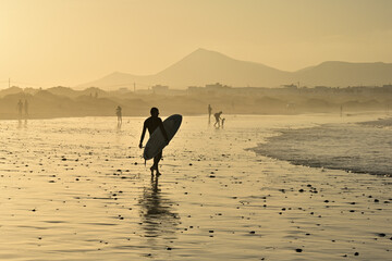 Ein Surf Boarder läuft durch den nassen Sand am Strand von Famara auf Lanzaroteund trägt dabei sein Board unter dem Arm, während im Hintergrund die Berge im Abendrot zu sehen sind - obrazy, fototapety, plakaty