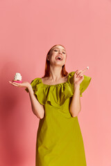 Portrait of young beautiful girl in cute dress posing with cupcake over pink background. Happy, positive vibe