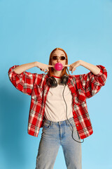 Fototapeta premium Portrait of young redhead girl in casual checkered shirt, sunglasses and headphones posing with bubblegum over blue background