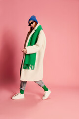 Portrait of stylish young girl in sunglasses, blue hat, green scarf and fur coat posing over pink background