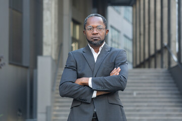 Serious and confident African American man in business suit looking at camera, businessman with crossed arms outside office building.