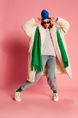 Portrait of young girl in sunglasses, blue hat, green scarf and fur coat posing over pink background. Stylish youth