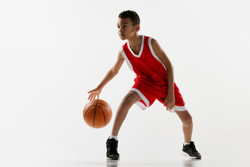 Naklejka premium Portrait of boy in red uniform training, playing basketball, dribbling over grey studio background. Motivated sportsman