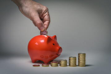 Hand steckt eine 1 Euro Münze zum sparen in ein Sparschwein mit anwachsenden Münzstapeln davor