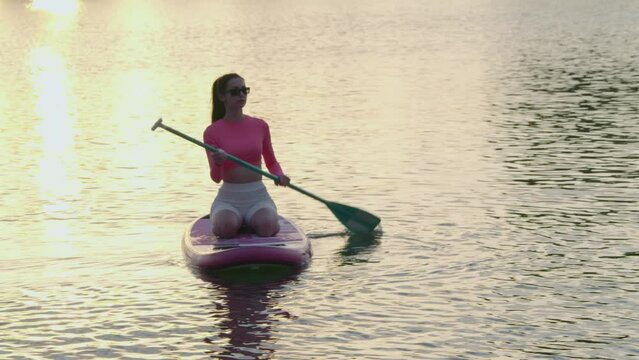 Healthy and fit woman floating on sup board with long paddle in hands. Caucasian young lady wearing sunglasses and sport clothes. Summer evening for outdoors workout. 