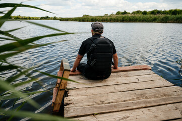 A man sits on the edge of a wooden bridge near a lake next to reeds.