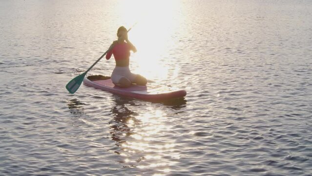 Young healthy woman wearing sport clothes swimming on sup board with long paddle and enjoying amazing sunset over lake. Concept of healthy and active lifestyle.
