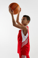 Portrait of boy in red uniform training, playing basketball over grey studio background. Concentration