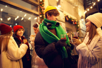 Happy young men and girls, friends walking at festive Christmas street and drinking mulled wine. Concept of holidays, winter, New Years fairs, friendship