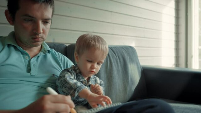 Little boy watches father drawing with marker in notepad. Man entertains curious toddler son sitting on comfortable sofa in living room