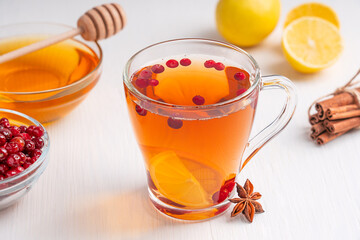 Homemade natural antioxidant hot tea healthy drink made with red cranberries, lemon citrus slices, cinnamon and sweet honey served in transparent glass cup on white wooden background with ingredients
