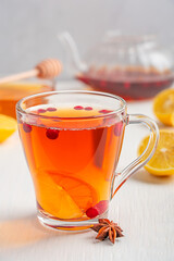 Natural antioxidant healthy hot tea drink made with cranberries, lemon citrus slices, anise and sweet honey served in transparent glass cup on white wooden table with ingredients and teapot at kitchen