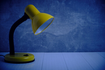 Bright yellow desk lamp, turned off, standing in dark against background concrete wall.