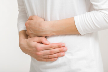 Young adult woman in white clothes touching painful belly with arms isolated on light gray background. Closeup. Side view. Girl suffering from stomach ache.