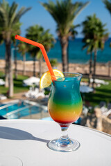 Colourful cold Rainbow Paradise cocktail drink served in glass at pool bar overlooking blue pool, sea and palm trees, relax and holidays at sea