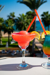 Colourful cold Strawberry daiquiri and Rainbow Paradise cocktails drinks served in glasses at outdoor cafe overlooking blue sea and palm trees, relax and holidays at sea