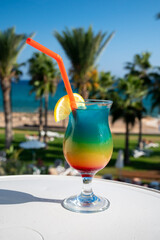 Colourful cold Rainbow Paradise cocktail drink served in glass at pool bar overlooking blue pool, sea and palm trees, relax and holidays at sea
