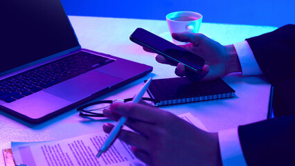 Hands of business person. Working desk with laptop, notebook, coffee, phone and documents in neon colored light