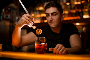 male bartender holds slice of dry citrus with tweezers over glass with cold cocktail