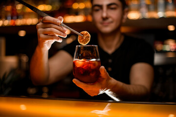 focus on hand of bartender with glass with cocktail garnishes it with slice of dry orange