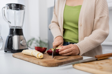 Cropped view of woman cutting ripe apple near banana and blurred blender in kitchen