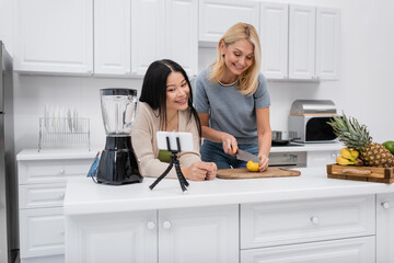 Multiethnic bloggers cutting lemon near smartphone on tripod in kitchen - Powered by Adobe
