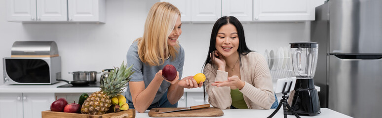 Smiling asian woman pointing at fruits near friend and smartphone on tripod in kitchen, banner
