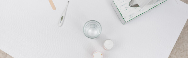 Top view of electronic thermometer near glass of water and pills on table at home, banner